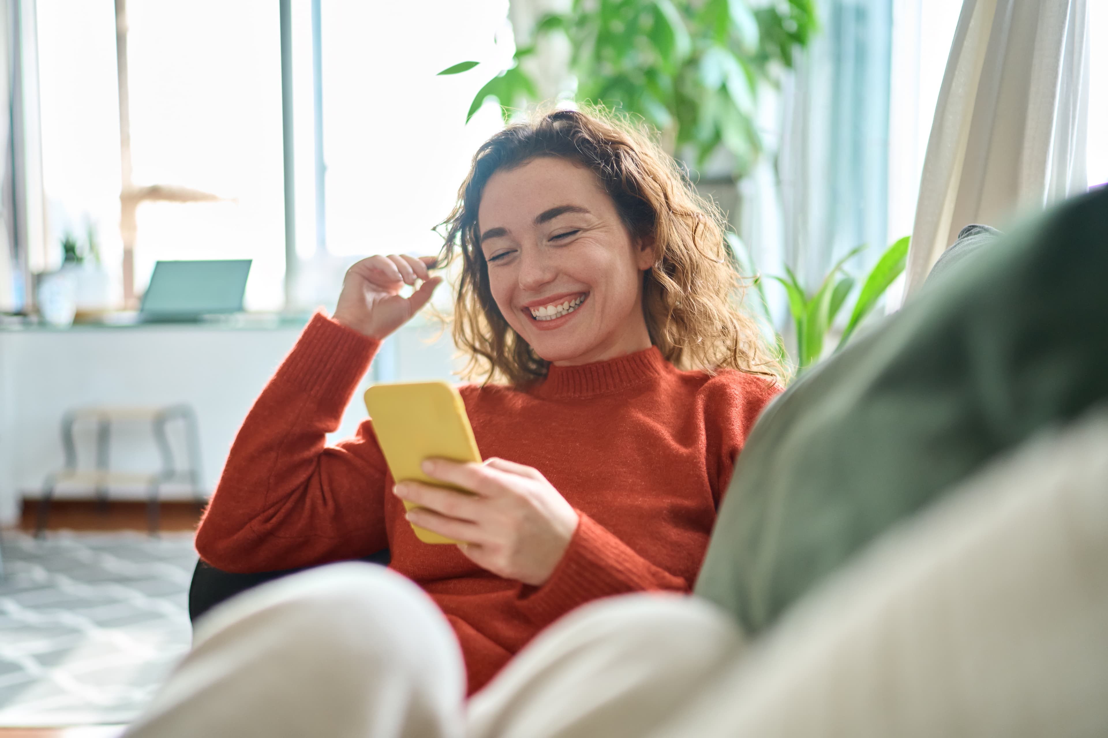 Woman laughing during a video call, seated on a modern armchair.